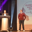 Der Preisträger der Kategorie "Junges Engagement"steht auf der Bühne. Links neben ihm hält jemand eine Rede an einem Pult. 