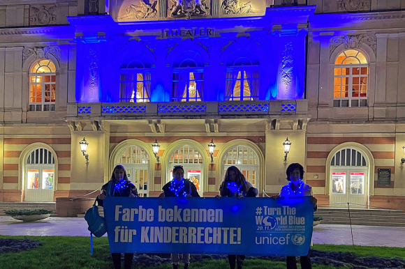 Ursula Grass, Astrid Kränzlein, Katharina Stegmaier und Margret Mergen (von links) halten vor dem Blau beleuchteten Theater einen Banner mit dem Text: "Farbe bekennen für Kinderrechte". Neben dem Text ist das Logo von unicef zu sehen.