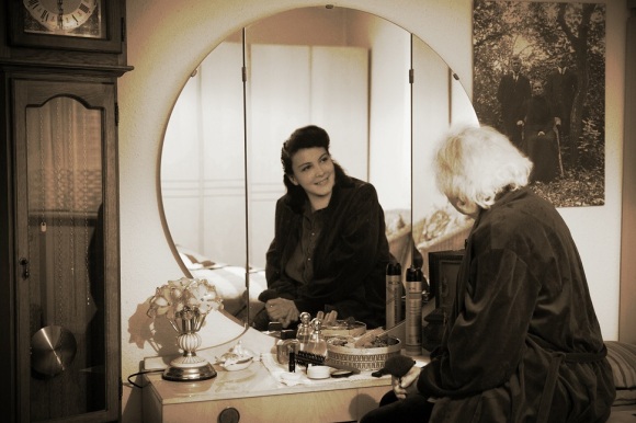 Eine ältere Frau mit grauen Haaren sitzt auf einem Hocker, schaut in den Spiegel und sieht eine junge Frau.