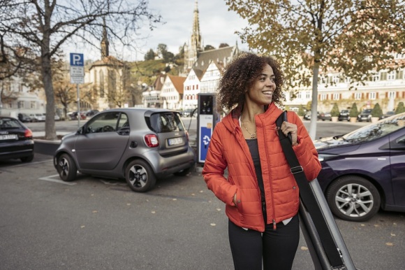 Eine lachende Frau steht vor mehreren Fahrzeugen. Eines davon ist ein E-Smart an einer Ladestation. 