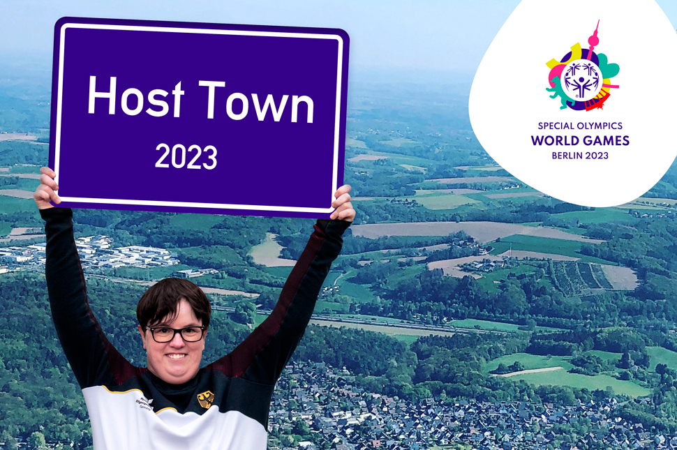 Juliana Rößler, Athletenrat SO Berlin, hält ein Schild auf dem steht: "Host Town 2023". Im Hintergrund eine Luftbildaufnahme. Rechts oben das Logo der Special Olympics.