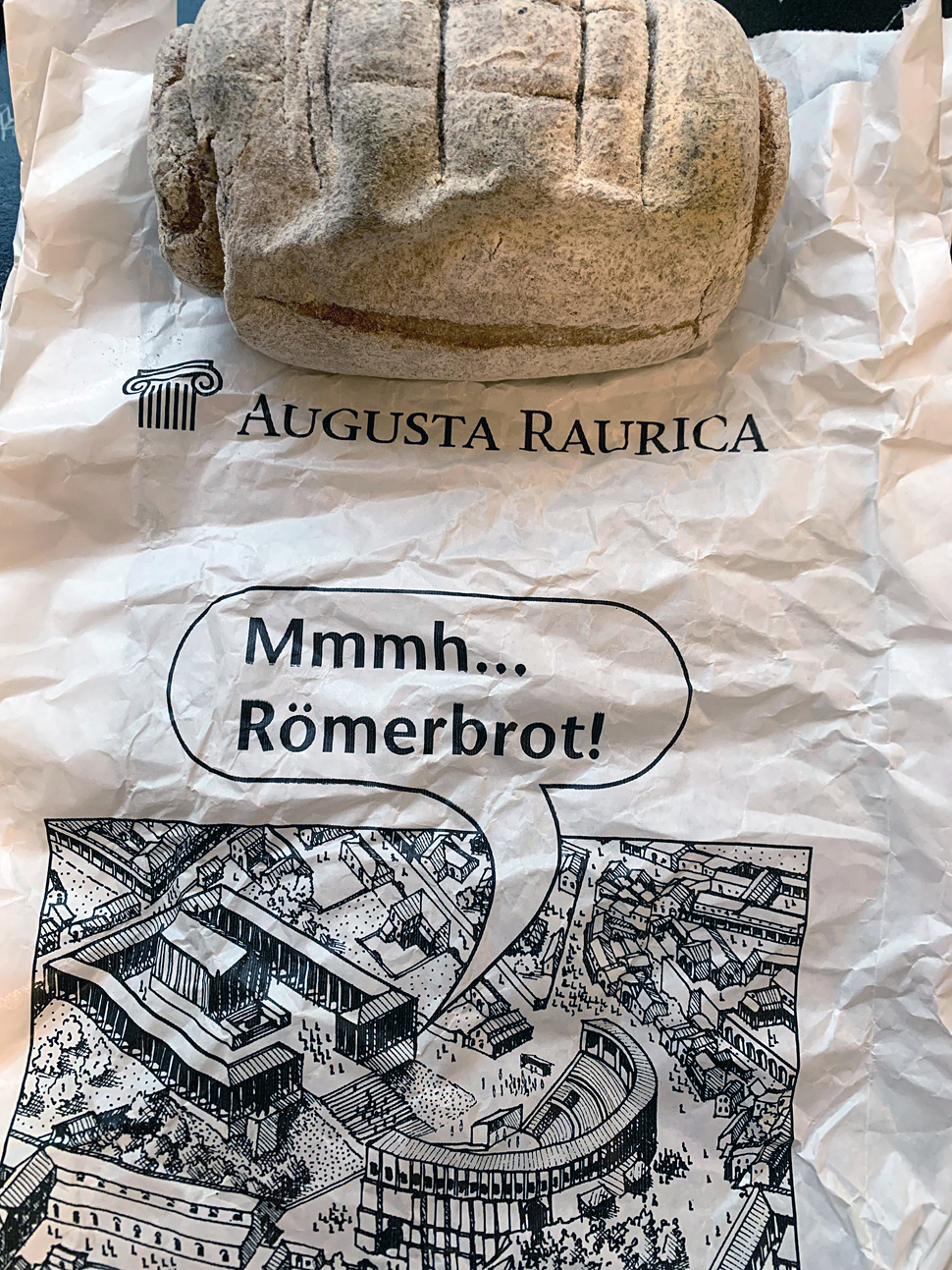 Zeichnung einer Römischen Stadt mit dem Text: "Augusta Raurica; Mmmh... Römerbrot!". Auf dem Papier liegt ein Stein