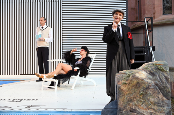 Szene aus dem Theaterstück "Was ihr wollt": Eine Frau liegt mit Bademantel und Badekleidung an einem Pool. Neben ihr steht ein Butler. Im Vordergrund eine Frau in Anzug.