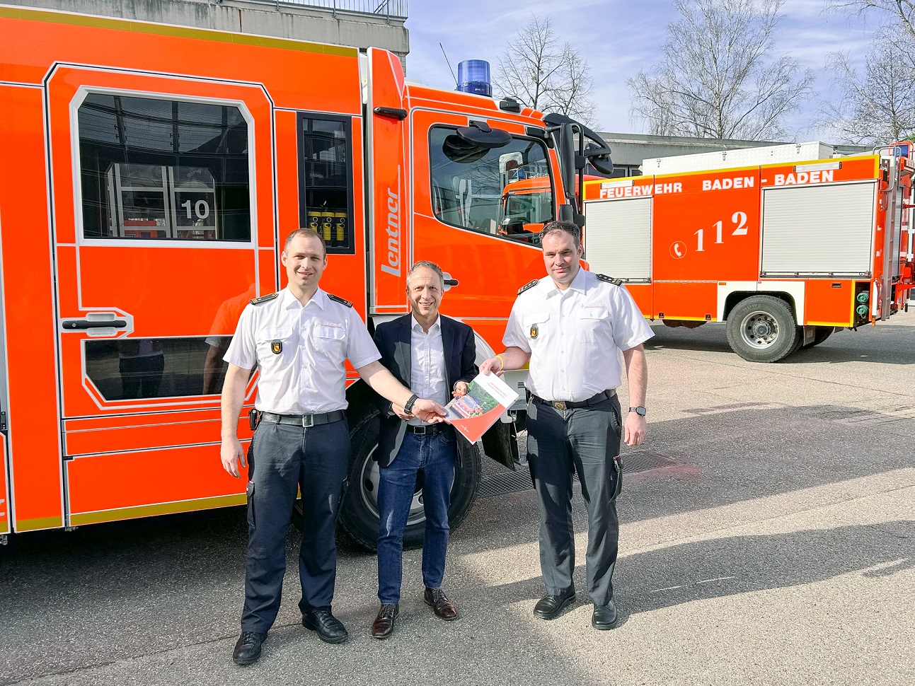 Bürgermeister Kaiser (Mitte) steht mit Kommandant Christian Pilardeaux (links) und dessen Stellvertreter Sascha Mundy (rechts) vor einem Feuerwehrauto und hält den Jahresbericht der Feuerwehr in den Händen. 