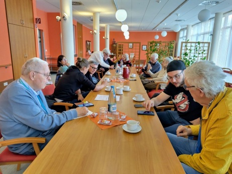 Seniorinnen und Senioren sitzen mit Jugendliche an einem Tisch und schauen auf Smartphones. 