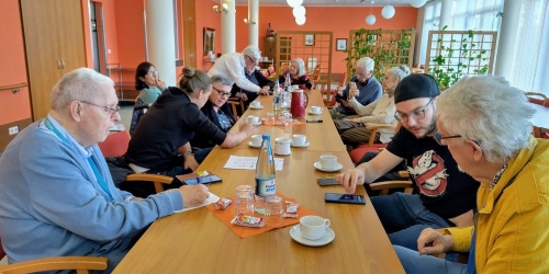 Seniorinnen und Senioren sitzen mit Jugendliche an einem Tisch und schauen auf Smartphones. 