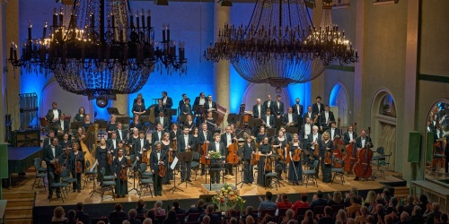 Ein Orchester steht auf einer Bühne vor Publikum. 