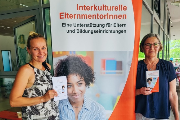 Zwei Frauen stehen vor einem Plakat mit der Aufschrift "Interkulturelle Elternmentoren"