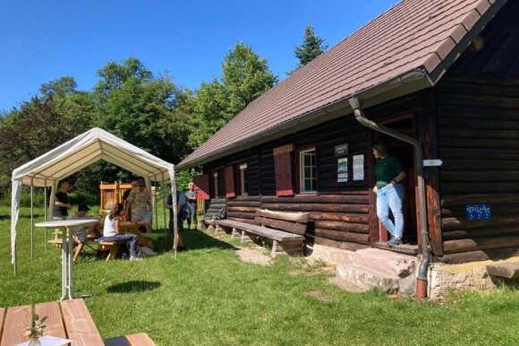 Vor einer dunklen Holzhütte steht ein weißes Pavillon, unter dem Menschen sitzen.