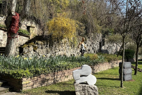 Auf einer grünen Wiese, vor einer niedrigen Steinmauer steht eine Steinskulptur.