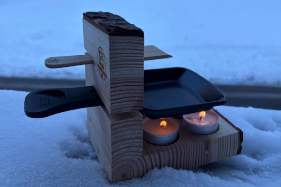 Ein mobiles Raclette Gerät mit Teelicht steht im Schnee. 