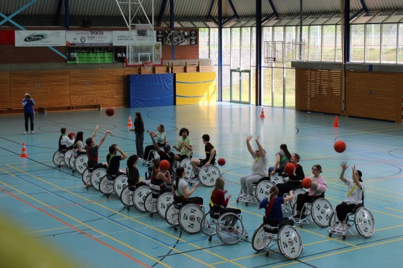 Sportlerinnen und Sportler in Rollstühlen werfen sich Basketbälle in einer Sporthalle zu. 