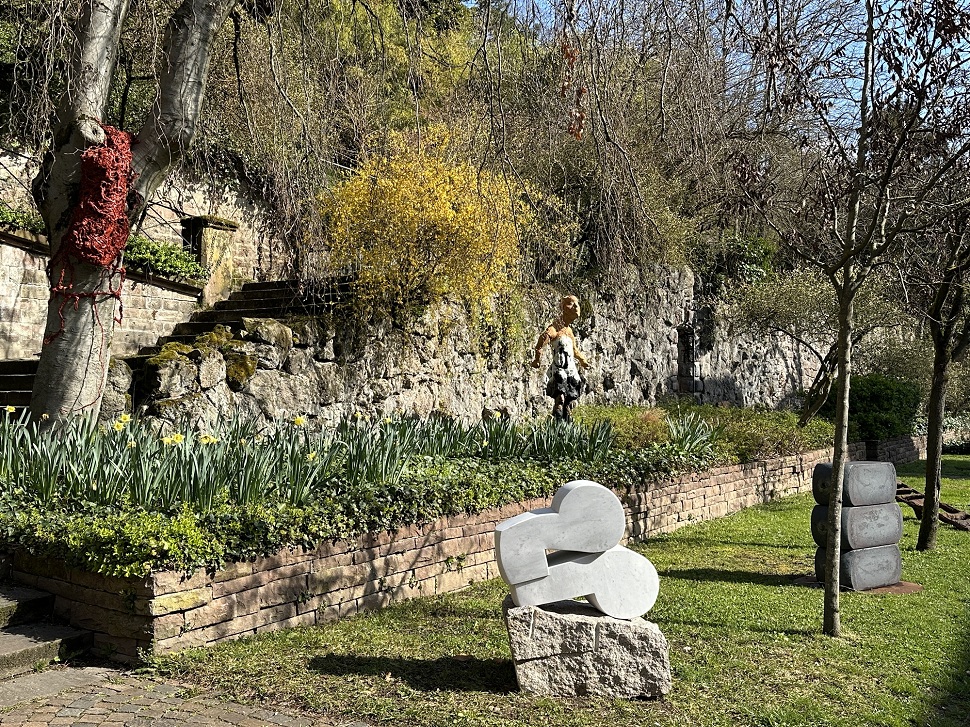 Auf einer grünen Wiese, vor einer niedrigen Steinmauer steht eine Steinskulptur.