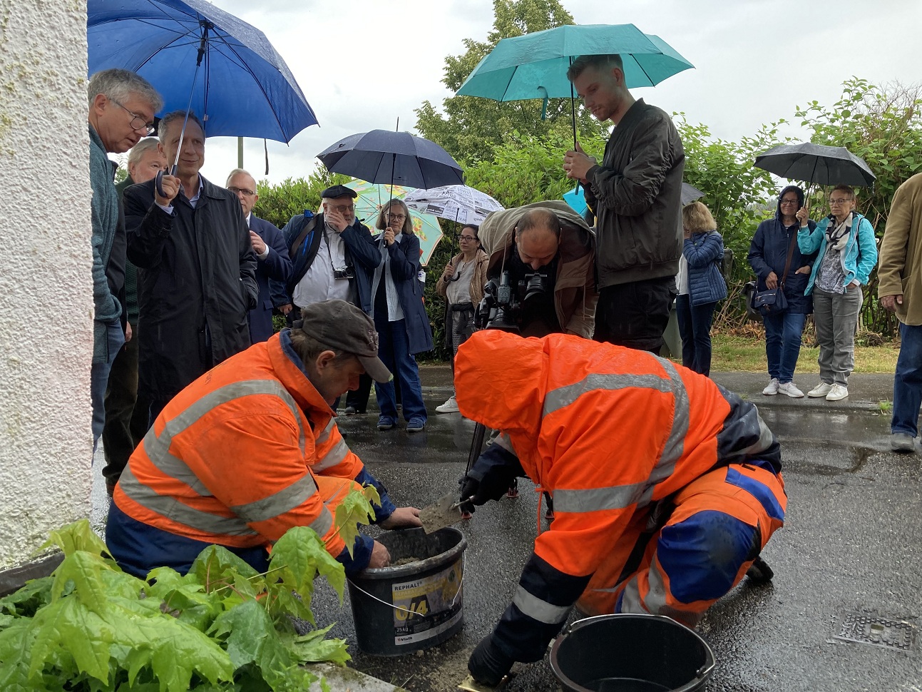 Zwei Männer lassen einen Stolperstein in den Asphalt ein. Mehrere Menschen mit Regenschirmen stehen drum herum. 