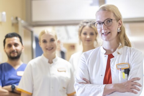 Eine Ärztin in weißem Kittel steht im Krankenhaus und verschränkt ihre Arme. Dahinter sind Pflegekräfte zu sehen.