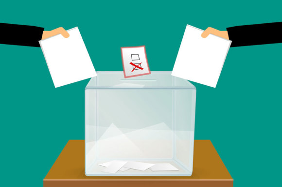 Grafik: Zwei Hände werfen weiße Blätter in eine Wahlurne. Über der Wahlurne ein Zettel mit einem Kreuz.