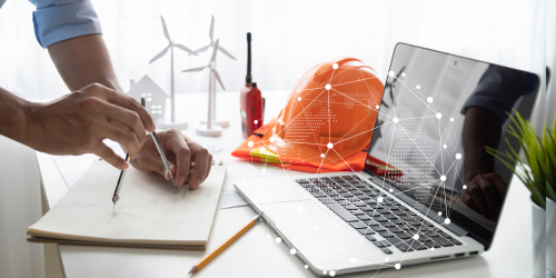 Männerhände zeichnen mit einem Zirkel einen Kreis auf Papier. Daneben steht ein Laptop und Helm.