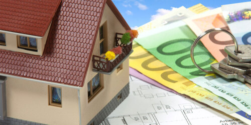 Ein Spielzeughaus steht auf Bauplänen, daneben Geldscheine und Hausschlüssel.