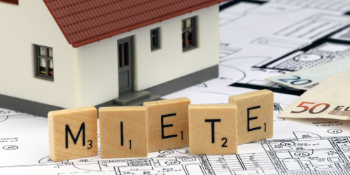 Spielzeughaus auf Bauplänen. Davor mit Holzklötzen das Wort Miete geschrieben. Daneben Geldscheine.