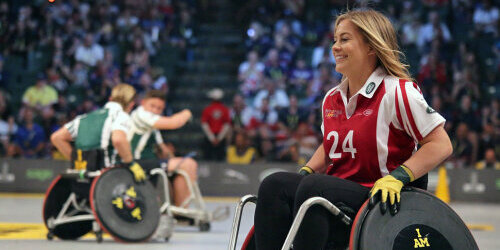 Frau im Rollstuhl in Sporttrikot in einer Halle. Im Hintergrund zwei Männer im Rollstuhl.