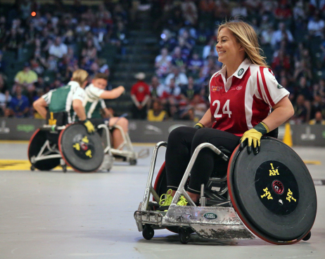 Frau im Rollstuhl in Sporttrikot in einer Halle. Im Hintergrund zwei Männer im Rollstuhl.