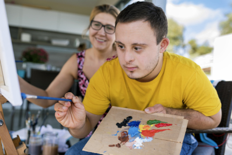 Teenager-Künstler mit Downsyndrom malt. Im Hintergrund sitzt eine Frau und beobachtet ihn.