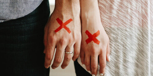 Hand von einem Mann und einer Frau. Der Mann trägt einen Ehering. Auf beiden Händen ist ein großes, rotes Kreuz zu sehen.