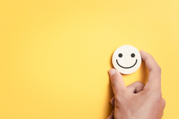 Eine Hand hält einen Smiley aus Holz vor einem gelben Hintergrund.