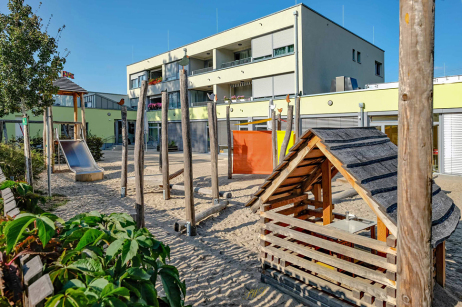 Außenansicht Gebäude mit Spielplatz. Spielplatz mit Rutsche, Balancierstange und Holzhütte