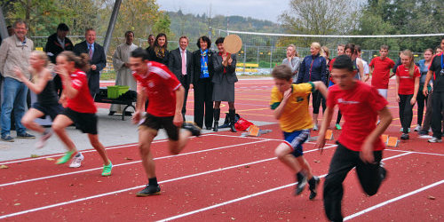 Mehrere Kinder sprinten auf der neuen Laufstrecke im Schulzentrum West. Im Hintergrund steht OB Mergen.
