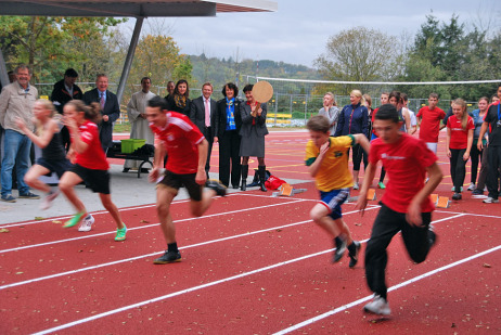 Mehrere Kinder sprinten auf der neuen Laufstrecke im Schulzentrum West. Im Hintergrund steht OB Mergen.