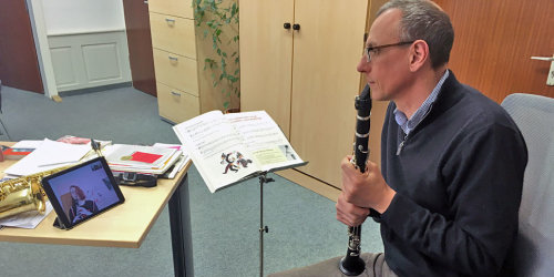 Musikschulleiter Ralf Eisler sitzt vor eine Tablet und spielt auf seiner Klarinette. In dem Tablet ist eine Musikschulerin, ebenfalls mit einer Klarinette, zu sehen.