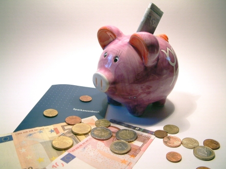 Ein Sparschwein in dem ein Geldschein steckt. Nebendran ein Sparkassenbuch Geldscheine und Münzen