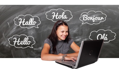 Jugendliche lächelt in die Kamera und sitzt mit Laptop vor einer Schultafel. Die Tafel ist mit "Hallo" in verschiedenen Sprachen beschriftet.