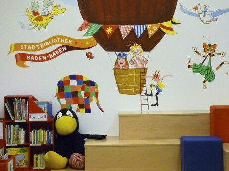 Treppe und bemalte Wand in der Kinderbibliothek. Auf der Treppe sitzt eine große Kuscheltierausgabe vom Raben Socke.
