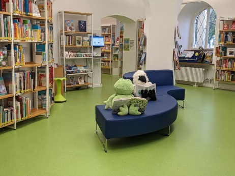 Blaues Sofa steht in der Kinderbibliothek. Auf dem Sofa sitzen zwei Kuscheltiere mit aufgeschlagenen Büchern.