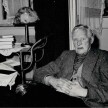 Otto Flake sitzt auf einem Sofa, die Hände ineinander verschränkt. Seitlich neben ihm ein Beistellwagen mit einer Lampe und einem Stapel Bücher. Davor auf einem runden Tisch liegen zwei Bücher und eine Brille 