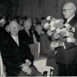 Festakt: Mehrere Menschen sitzen in Stuhlreihen. In vorderster Reihe sitzt Otto Flake. OB Schlapper steht vor ihm und hält einen Blumenstrauß für Flake in den Händen