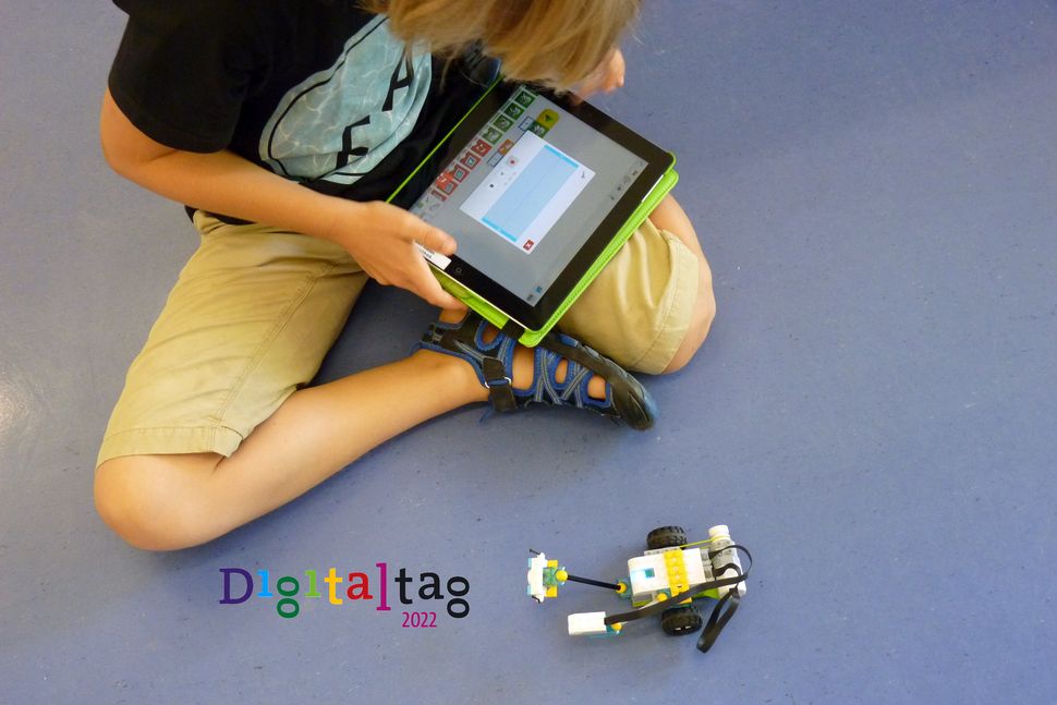Junger Mann mit iPad und Lego-Fahrzeug auf dem Boden sitzend. Daneben Logo des Digitaltags.
