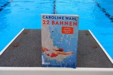 Buch steht auf Startblock im Schwimmbad, dahinter Wasser.
