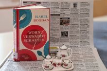 Ein aufgestelltes Buch mit dem Titel "Wohnverwandschaften" und Puppengeschirr vor einer Zeitung mit Wohnungsanzeigen.