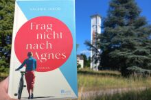 Im Hintergrund eine moderne Kirche. Davor ein Buchcover mit einer Frau, die ein Fahrrad schiebt.