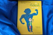 Gelbes Kinderbuch liegt auf blauem Stoff. Das Cover zeigt einen kleinen Junge und dahinter eine große Fantasiegestalt, die ihren Bizeps zeigen.