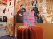 Buch über Komponistinnen in rosa vor Hintergrund einer Museumswand mit ebenfalls Komponistinnen.