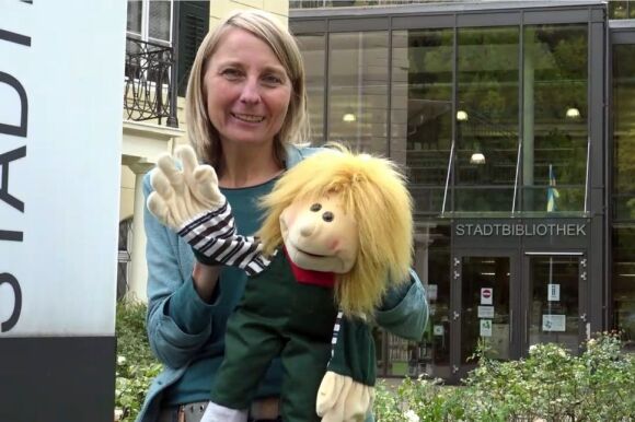 Frau mit Handpuppe vor dem Eingang der Stadtbibliothek mit Glasfront.