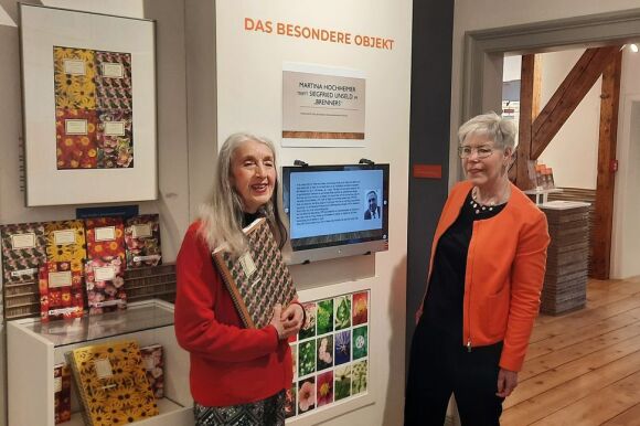 Zwei Frauen vor einer Ausstellungswand mit Rosenbüchern. Die Künstlerin hält einen großen Bildband vor ihrem Körper.