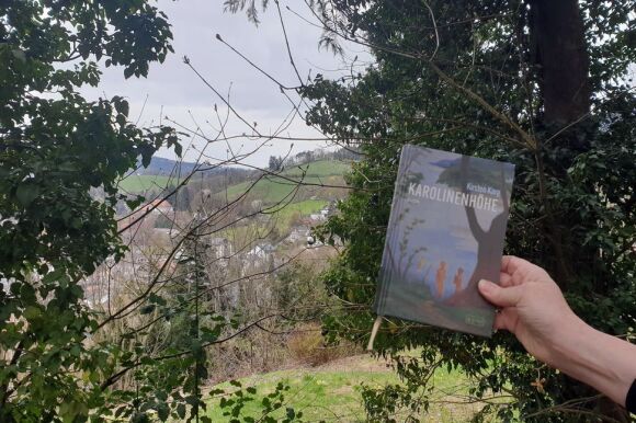 Auf dem Cover des Buches Karolinenhöhe sieht man zwei Menschen, die in der Natur unter einem Baum stehen (künstlerische Darstellung). Jenseits des Balkons sieht man eine hügelige Landschaft in Natura.