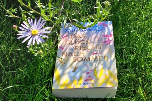 Buch in Pastellfarben liegt im Gras neben einer Blume.