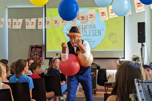 Akteur in Pose mit Luftballons vor Kindern auf Stühlen.