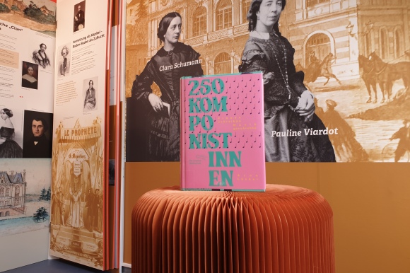 Buch über Komponistinnen in rosa vor Hintergrund einer Museumswand mit ebenfalls Komponistinnen.